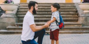 A father and his son outside at school. The child wears a backpack and father kneels down to speak with the boy